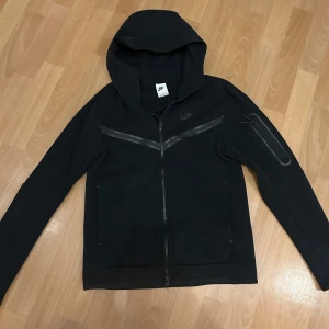 Svart Nike hoodie Tech Fleece, stl S - Svart Nike Tech Fleece hoodie med hel dragkedja och huva. Snygga svarta detaljer i form av tejpade linjer över bröstet och en ficka med dragkedja på ärmen. Mjuk och sportig känsla, perfekt för chill eller träning.