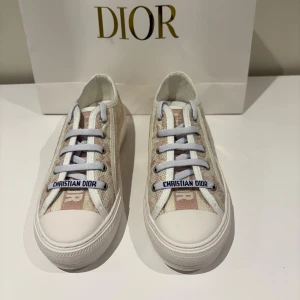 Beige och vita sneakers från Dior - Snygga sneakers från Dior i rosa/beige och vitt med diskret mönster och tydlig Christian Dior logga på snörningen. Skorna har vit platt sula med svart stjärndetalj undertill. Säljer dem åt min syster kom med bud!