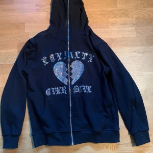 Trotherz rhinestone hoodie - Svart hoodie med dragkedja och huva. På framsidan finns ett stort tryck med texten 'Loyalty Over Love' och ett glittrigt hjärta i rhinestone. Skriv vid minsta lilla fundering!