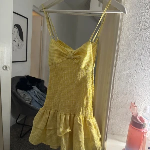 Gul rutig kortklänning med rosett YAMZ - Supersöt gul kortklänning med rutigt mönster, smockad midja och volangkant från YAMZ. Klänningen har smala axelband, stor rosett framtill och öppen rygg. Perfekt för varma dagar och ger en riktigt somrig vibe.