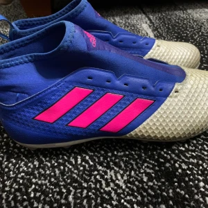 Blå och vita adidas fotbollsskor - Säljer ett par blåa adidas fotbollsskor med rosa ränder och vita dobbar. Skorna har en stickad ovandel och högre skaft för extra stöd. Perfekta för dig som vill sticka ut på planen med snygg design och bra grepp.