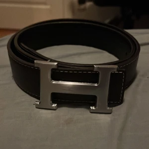 Svart H bälte - Snyggt svart bälte från Herme med klassiskt stort H-spänne i silverfärgad metall. Bältet är gjort i äkta skinn med tydliga sömmar längs kanterna och har en stilren, tidlös design som passar till jeans eller kostymbyxor. Knappt använd max 2 gånger