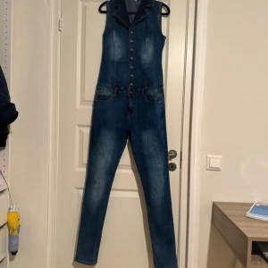 Ärmlös jeans jumpsuit med knappar - Snygg ärmlös jeans jumpsuit i klassisk blå denim med krage och knappar framtill. Tight passform med raka ben, fickor fram och bak samt coola sömdetaljer. Perfekt för dig som vill sticka ut med en unik denimlook.