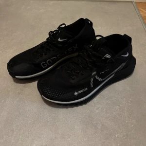 Nike React Pegasus Trail 4 Gore Tex strl 42,5 - Svarta Nike sneakers med Gore-Tex-material, perfekt för regniga dagar. Snygg design med vita detaljer och reflekterande Swoosh på sidan. Skorna har snörning, slitstark sula och är tillverkade i syntetmaterial. Modellnummer DJ7926-001.