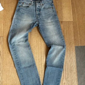 Levi's 501 ljusblå jeans W28 L32 - Klassiska Levi's 501 jeans i ljusblå tvätt med raka ben och femficksmodell. Jeansen har knappgylf, snygga slitningar och den ikoniska läderpatchen bak. Perfekta för dig som gillar en tidlös och avslappnad stil.