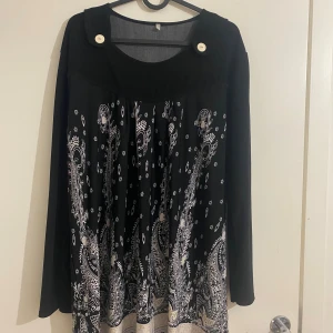 Svart långtröja - Svart llångtröja  med vit paisleyinspirerat mönster och dekorativa knappar vid axlarna. Klänningen har en lös passform och rund halsringning. Materialet känns mjukt och faller snyggt. Perfekt för dig som gillar unika detaljer och stilrena kontraster.