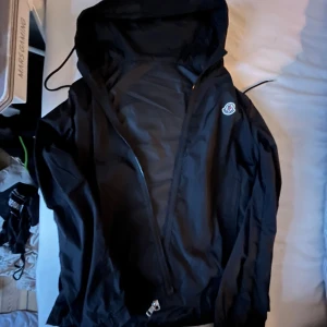 Moncler windbreaker - Säljer en svart windbreaker från Moncler med huva och dragkedja framtill. Jackan har Moncler-logga på bröstet och en clean, sportig look. Perfekt för dig som vill ha en stilren jacka med premiumkänsla. Skön till höst starten