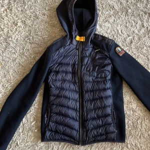 Mörkblå pufferjacka från Parajumpers - Säljer en mörkblå pufferjacka från Parajumpers med huva och coolt patch på ärmen. Jackan har quiltad framsida och rygg, dragkedja framtill och fickor med dragkedja. Ärmarna är ribbade och jackan har en sportig vibe. Perfekt för kyliga dagar!