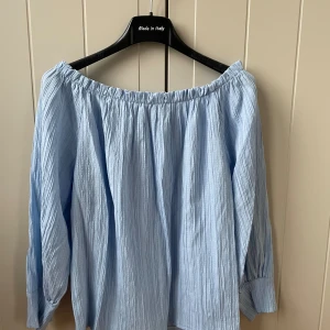 Ljusblå offshoulder blus New Collection - Superfin ljusblå blus från New Collection med offshoulder-modell och långa ärmar. Blusen har en krinklad struktur och är luftig och flowy, perfekt för varmare dagar. Resår upptill för snygg passform över axlarna.