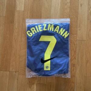 Atlético Madrid Griezmann Nike tröja - Snygg blå Atlético Madrid matchtröja med Griezmann och nummer 7 i gult på ryggen. Nike Dri-Fit material, officiell klubbmärkning och sponsortryck i gult. Coola blixtdetaljer på sidorna och v-ringad hals. Perfekt för dig som älskar fotboll och Griezmann.