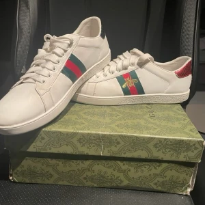 Gucci vita sneakers med röd/grön detalj - Säljer ett par vita sneakers från Gucci i skinn med klassiska rödgröna ränder på sidorna och broderad guldbi. Baksidan har röd och grön detalj i ormskinnsmönster. Skorna har snörning och platt sula, perfekt för dig som vill sticka ut med lyxig streetstyle.