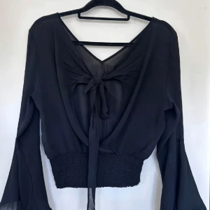Svart blus med knyt och vida ärmar - Säljer en svart blus i tunn, luftig polyester med v-ringning och knyt baktill. Blusen har vida, långa ärmar och smockad midja som ger en snygg passform. Perfekt för dig som gillar trendiga detaljer och en lite bohemisk vibe.