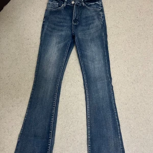 Blå bootcut jeans med detaljerade bakfickor - Snygga blå jeans med bootcut passform och coola detaljerade bakfickor med knappar. Jeansen har klassisk femficksdesign, lätt slitning framtill och är tillverkade i mjukt denimtyg. Perfekta för dig som gillar en retroinspirerad look. Använd 1-2 gånger bara!