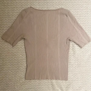 Beige ribbad topp med djup rygg - Säljer en beige ribbad topp med korta ärmar och snyggt djup rygg. Toppen har en figurnära passform och är tillverkad i ett stretchigt material som sitter skönt på kroppen. Perfekt att styla med jeans eller kjol för en clean look.