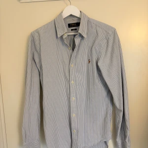 Randig skjorta från Polo Ralph Lauren - Säljer en klassisk blåvit randig skjorta från Polo Ralph Lauren i slim fit. Skjortan har långa ärmar, knappar framtill och den ikoniska broderade loggan på bröstet. Tillverkad i mjuk bomull och passar perfekt för en clean och stilren look.