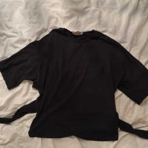 Svart T-shirt/topp med knyt i ryggen - Säljer en svart topp från Nelly med korta ärmar och snygg knytning i ryggen som ger en öppen rygg-effekt. Toppen har en loose passform upptill och blir tightare i midjan tack vare knytbanden. Perfekt till jeans eller kjol för en trendig look.💕💕