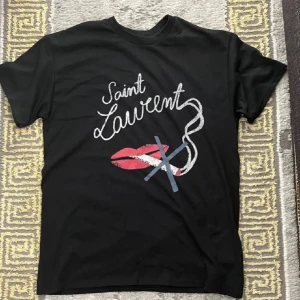 Yves Saint Lauren ”no smoking” t-shirt - Svart t-shirt från Saint Laurent med coolt grafiskt tryck framtill i vitt, rosa och blått. Inga tags Trycket har handskriven text och ett motiv med läppar och linjer. Klassisk rund hals och korta ärmar. Första bilden är tagen av en vän inte den som jag säljer är bara samma modell. Perfekt för dig som gillar statement-plagg och designer.