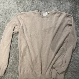 Beige stickad tröja från Bläck - En stilren beige stickad tröja från Bläck i storlek L. Tröjan har rund halsringning, ribbade muddar vid ärmslut och nederkant samt långärmad passform. Perfekt för lager-på-lager och enkel att matcha med jeans eller chinos.