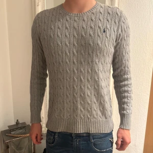 Grå kabelstickad tröja Polo Ralph Lauren - Kabelstickad grå tröja från Polo Ralph Lauren med klassisk rund halsringning och broderad marinblå logga på bröstet. Tröjan sitter som en M. Modellen på bilden är 183 cm. Den passar även folk som har storlek S.