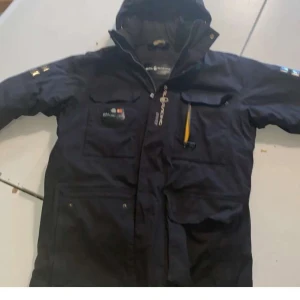 Svart vinterjacka Sail Racing - Svart vinterjacka från Sail Racing med huva, flera praktiska fickor och dragkedja framtill. Jackan har logga på bröstet och ryggen samt detaljer i gult. Perfekt för kalla dagar med sitt vadderade foder och sportiga look.