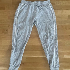 Grå mjukisbyxor från Gina Tricot L - Grå mjukisbyxor från Gina Tricot i storlek L, tillverkade i mjuk bomull. Byxorna har vita ränder längs sidorna, resår och snörning i midjan samt muddar vid bensluten. Perfekta för en chill och avslappnad stil.