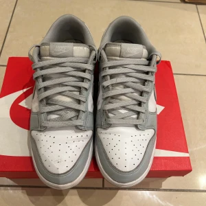 Nike Dunk Low vit/grå sneakers - Nike Dunk Low sneakers i vitt och ljusgrått skinn med klassisk Swoosh på sidorna. Lågt skaft, perforerad tå och matchande grå snörning. Snygg och clean siluett som passar till det mesta. Kommer med originalkartong.