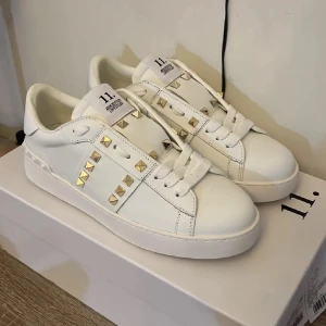 Valentino Garavani vita sneakers med nitar - Snygga vita sneakers från Valentino Garavani med ikoniska guldiga nitar på sidorna och tungan. Skorna har rund tå, platt sula och är gjorda i skinn. Klassisk snörning och diskret logga på plösen. Perfekta för dig som vill sticka ut med stilrena detaljer.
