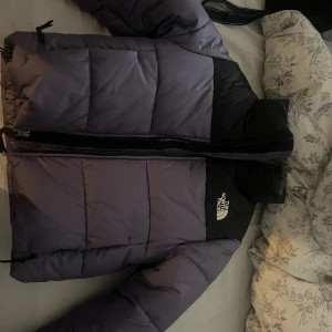 Lila och svart pufferjacka från The North Face - Säljer en snygg pufferjacka från The North Face i lila och svart. Jackan har hög krage, dragkedja framtill och logga på bröstet. Den är quiltad och har en loose passform, perfekt för kalla dagar. Materialet är syntet och dun för extra värme.