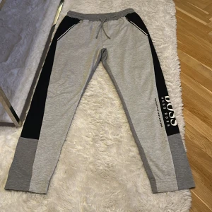 Grå mjukisbyxor Hugo Boss slim fit - Snygga grå mjukisbyxor från Hugo Boss med svarta paneler längs sidorna och vit logga på ena benet. Byxorna har snörning i midjan, slim fit-passform och är tillverkade i mjukt material. Perfekta för chill eller träning. Passar till S oxå.