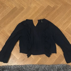 Svart blus med volanger från Forever 21 - Säljer en svart blus från Forever 21 med långa ärmar och snygga volangdetaljer framtill. Blusen har en v-ringad hals och är tillverkad i ett lätt, luftigt material som ger en skön känsla. Perfekt för dig som gillar stilrena och trendiga plagg.