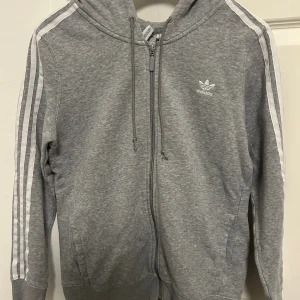 Grå Adidas hoodie med dragkedja - Grå hoodie från Adidas med klassiska vita ränder längs ärmarna och logga på bröstet. Tröjan har huva med snörning, dragkedja framtill och två fickor. Perfekt för en sportig och avslappnad stil.