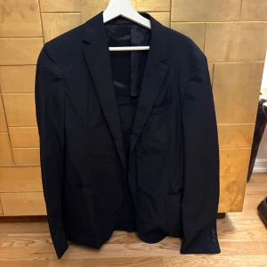 Svart kavaj från Prada - Säljer en svart kavaj från Prada med klassisk passform och enkla knappar vid ärmslutet. Kavajen har slät yta, smala slag och innerficka med tydlig Prada-logga. Perfekt för dig som vill ha en stilren och exklusiv look.