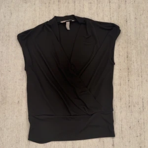 Svart omlottopp från H&M - Säljer en svart omlottopp från H&M med ärmlös design och bred linning nedtill. Toppen har en snygg omlott-look framtill och är gjord i mjukt, stretchigt material som sitter skönt mot kroppen. Perfekt för dig som gillar stilrena och enkla plagg.