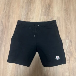 Svarta shorts från Moncler med snörning - Svarta shorts från Moncler med elastisk midja och snörning. Klassisk design med två sidofickor och Moncler-logga broderad på vänster ben. Mjukt bomullsmaterial som är skönt för sommardagar. Perfekta för en chill och stilren look.