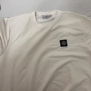 Vit sweatshirt från Stone Island - Vit långärmad sweatshirt från Stone Island med klassisk rund hals och ribbade muddar. Tröjan har en svart patch med logga på bröstet och är gjord i mjuk bomull. Perfekt för dig som gillar clean och stilren streetwear.