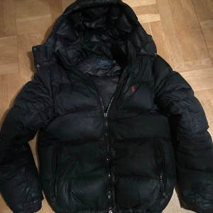 Svart dunjacka Polo Ralph Lauren - Svart dunjacka från Polo Ralph Lauren med huva och dragkedja. Jackan har en klassisk pufferstil, två fickor med dragkedja och ikonisk röd logga på bröstet. Perfekt för kalla dagar och riktigt snygg till streetwear. Finns ett hål vid armen och en del av dragkedjan har gått av funkar fortfarande utmärkt 