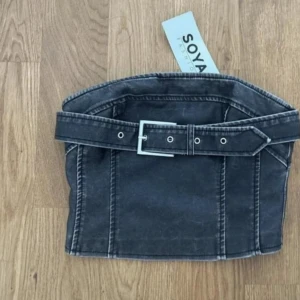 Svart denimkjol med bälte från Soya - Cool svart denimkjol från Soya med markerade sömmar och ett brett bälte i midjan. Top har en kort och rak modell som ger en edgy vibe. Perfekt för dig som gillar streetstyle och vill sticka ut med detaljer.den har en dragkedja där bak på ryggen.Onavänd! 