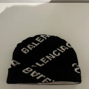 Svart mössa från Balenciaga - Snygg svart mössa från Balenciaga med vit logotyp i diagonal design över hela mössan. Stickad i mjukt material och har uppvikt kant. Perfekt accessoar för att lyfta din streetstyle.