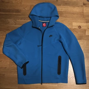 Blå Nike vindjacka med huva - Säljer en blå Nike vindjacka med huva och svarta detaljer. Jackan har hel dragkedja framtill, två fickor med svarta dragkedjor och Nike-logga på bröstet. Perfekt för dig som gillar sportig stil och vill ha något som sticker ut. Den är helt ny köpte den förra året tåg på mig den en gång sen så hade jag den i min garderob. Den är äkta jag har kvittot till den som behöver 