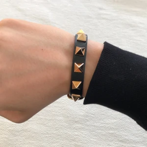 Svart armband med guldfärgade nitar - Snyggt svart valentino armband med fyrkantiga guldfärgade nitar och spänne. Armbandet är gjort i skinn och har justerbar längd med flera hål för perfekt passform. Perfekt accessoar för att ge din outfit en edgy touch. Passar till allt🤩✅