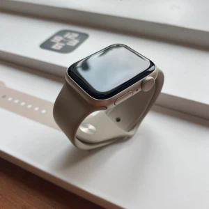 Apple Watch SE 2nd gen 40mm - Säljer en knappt använt apple watch i färgen starlight. Batteri hälsan är 100% och skickar med två skärmskydd och en extra band i färgen champagne som inte finns med i bilden.