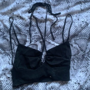 Svart croppad linnetopp från Zara - Säljer en svart croppad linnetopp med smala axelband och snygg snörning framtill som går att knyta i nacken. Toppen har en ribbad, elastisk nederdel och ärmlös design som ger en cool och trendig look. Perfekt att styla med höga jeans eller kjol. Toppen är använd 2 gånger och är i ny skick!😇