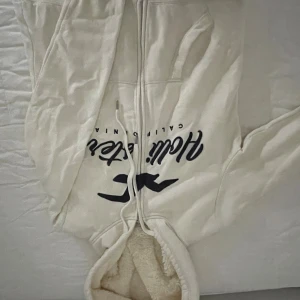 Vit hoodie med teddyfoder från Hollister.  - Mysig vit hoodie från Hollister med teddyfoder i hela insidan och huva. Dragkedja framtill, snörning vid huvan och två fickor. Stor Hollister-logga i mörkblått på bröstet. Är köpt från sellpy men fortfarande i bra skick( inga större slitningar förutom lite nopprig o har en knappt synlig fläck på ena ärmen). Snygg och bekävm passform( är igentligen i storlek m, men passar även xs och s).🤍🤍