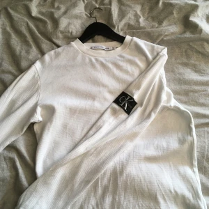 Vit långärmad tröja från Calvin Klein - Säljer en stilren vit långärmad tröja från Calvin Klein med diskret ribbad struktur och svart CK-logga på ärmen. Perfekt för dig som gillar enkel och clean stil. Tröjan har rund halsringning och är riktigt skön att bära. Ops! Litet hål där bak