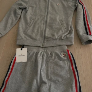 Grå zip tracksuit från Moncler - Grå hoodie från Moncler med hel dragkedja och ribbad krage. Snygga röda, vita och marinblå ränder längs ärmarna och Moncler-logga på bröstet. Tillverkad i mjuk bomull, perfekt för en sportig och avslappnad look. Passar S/m
