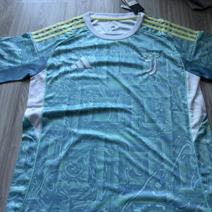 Juventus bortaställ 25/26 (Rabatt) - Juventus fotbollströja från Adidas i blått och vitt med unikt abstrakt mönster. Tröjan har gula ränder på axlarna, broderad Juventus-logga med tre stjärnor och Adidas-märke på bröstet. Tillverkad i lätt och ventilerande polyester, perfekt för match eller träning.