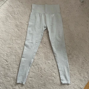 Grå Gymshark träningsbyxor - Snygga ljusgrå leggings från Gymshark med diskret mönstrad struktur längs benen. Stretchigt material som sitter tight och bekvämt, perfekt för träning. Gymshark-logga framtill vid midjan. S-XS