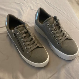 Olivgröna sneakers med sido dragkedja  - Snygga olivgröna sneakers med en silvrig detalj på hälen. Skorna har snörning framtill och en dragkedja på sidan för extra stilpoäng. Perfekta för dig som gillar en clean look. (Original pris 549) 