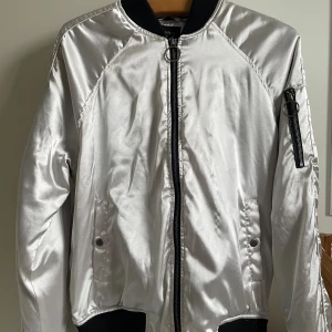 Silver bomberjacka från Forever 21 - Snygg silverfärgad bomberjacka från Forever 21 med svart ribbad krage, muddar och nederkant. Jackan har dragkedja framtill, fickor med tryckknapp och en extra ficka med dragkedja på ärmen. Tillverkad i glansig polyester. Passar båda kön. 