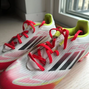 Adidas F50 vita fotbollsskor - Säljer ett par Adidas F50 fotbollsskor i vitt med svarta och röda detaljer, samt neongula inslag. Skorna har röda snören, platt sula med dobbar och klassiska Adidas-ränder på sidan. Ovandelen är i syntetmaterial och insidan är fodrad i neongult.